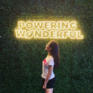 May include: A yellow neon sign mounted on a green wall displaying the text POWERING WONDERFUL in a bold, sans-serif font. The letter O in POWERING features a leaf-shaped cutout, and the letter O in WONDERFUL contains a lightning bolt symbol.