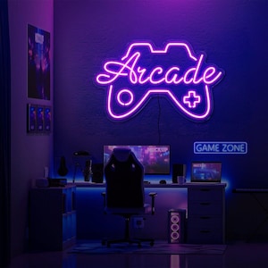 May include: A purple neon sign shaped like a game controller with the word Arcade in cursive script. Below, a desk setup features a computer monitor, a gaming chair, a laptop, and a smaller rectangular neon sign displaying the text GAME ZONE.