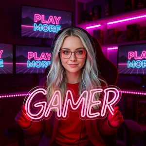 May include: A person with long light-colored hair and glasses wearing a red long-sleeve shirt, holding a pink neon sign that reads GAMER. The background features multiple computer monitors displaying the text PLAY MORE in neon pink and blue.