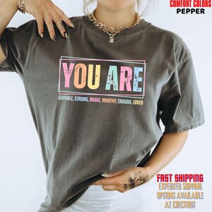 May include: A pepper-colored short-sleeve t-shirt featuring the text YOU ARE in large, multi-colored block letters inside a rectangular frame, with the words CAPABLE, STRONG, BRAVE, WORTHY, ENOUGH, LOVED printed in smaller text underneath.