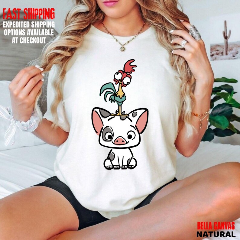 May include: A white Bella Canvas natural t-shirt featuring a cartoon illustration of a pig with a rooster on top. The pig is white with black spots and pink ears. The rooster is yellow, blue, and red. The shirt has the words "FAST SHIPPING" in red.