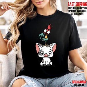 May include: Black t-shirt featuring a cartoon illustration of a pig with a rooster perched on top. The pig is white with black spots and pink ears. The rooster has red, blue, and yellow accents. The shirt has the text "BELLA CANVAS BLACK".