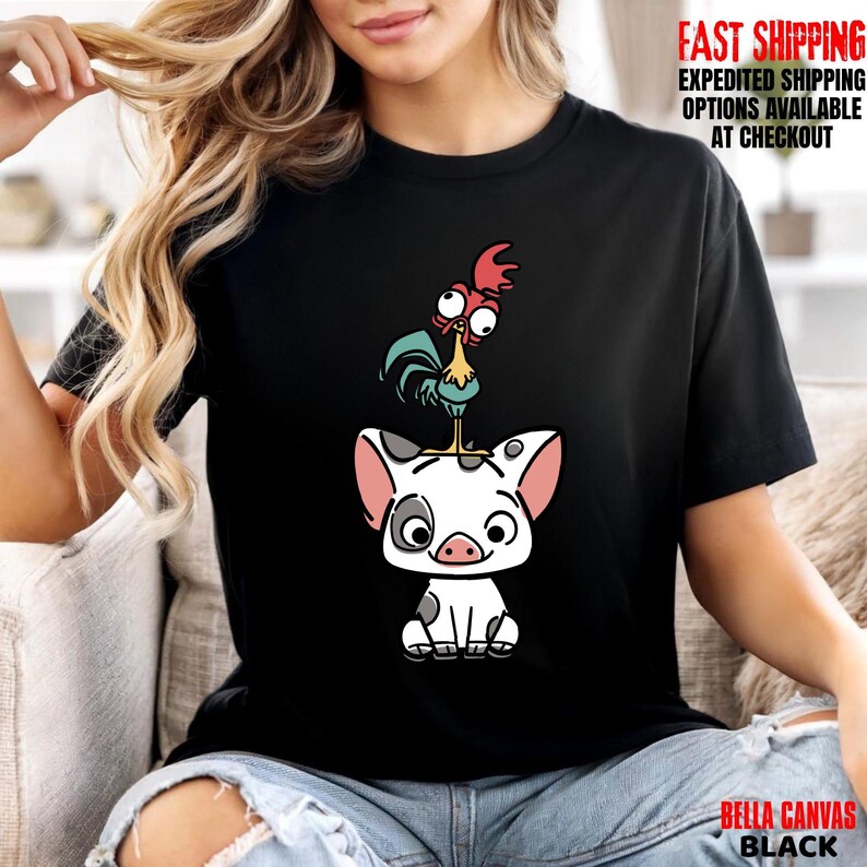 May include: Black t-shirt featuring a cartoon illustration of a pig with a rooster perched on top. The pig is white with black spots and pink ears. The rooster has red, blue, and yellow accents. The shirt has the text "BELLA CANVAS BLACK".