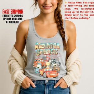 Puede incluir: Una camiseta sin mangas gris claro con un gr&aacute;fico colorido de la ciudad de Radiador Springs y varios personajes de coches de dibujos animados. El texto "Radiator Springs" se muestra encima de los coches. La camiseta sin mangas es sin mangas y tiene cuello redondo.
