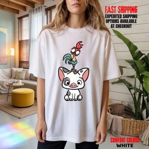 May include: White t-shirt featuring a cartoon illustration of a pig with a rooster on its head. The pig is white with black spots and pink accents. The rooster is blue, red, and yellow. The shirt also has the text "COMFORT COLORS WHITE".