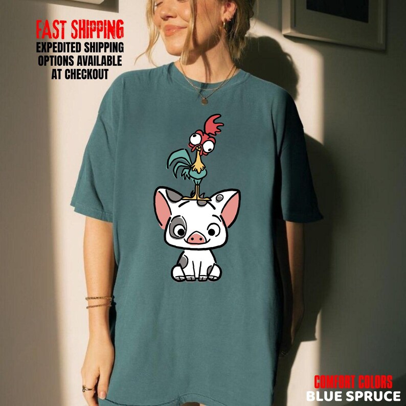 May include: Blue spruce t-shirt featuring a cartoon pig with a rooster on its head. The pig is white with black spots and pink ears. The rooster is red, blue, and green. The shirt has the text "COMFORT COLORS BLUE SPRUCE".
