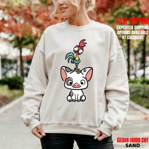 May include: Sand-colored sweatshirt featuring a cartoon illustration of a pig with a rooster on top. The pig is white with black spots and pink ears. The rooster is blue, yellow, and red. The sweatshirt has the text "GILDAN 18000 SWT SAND".