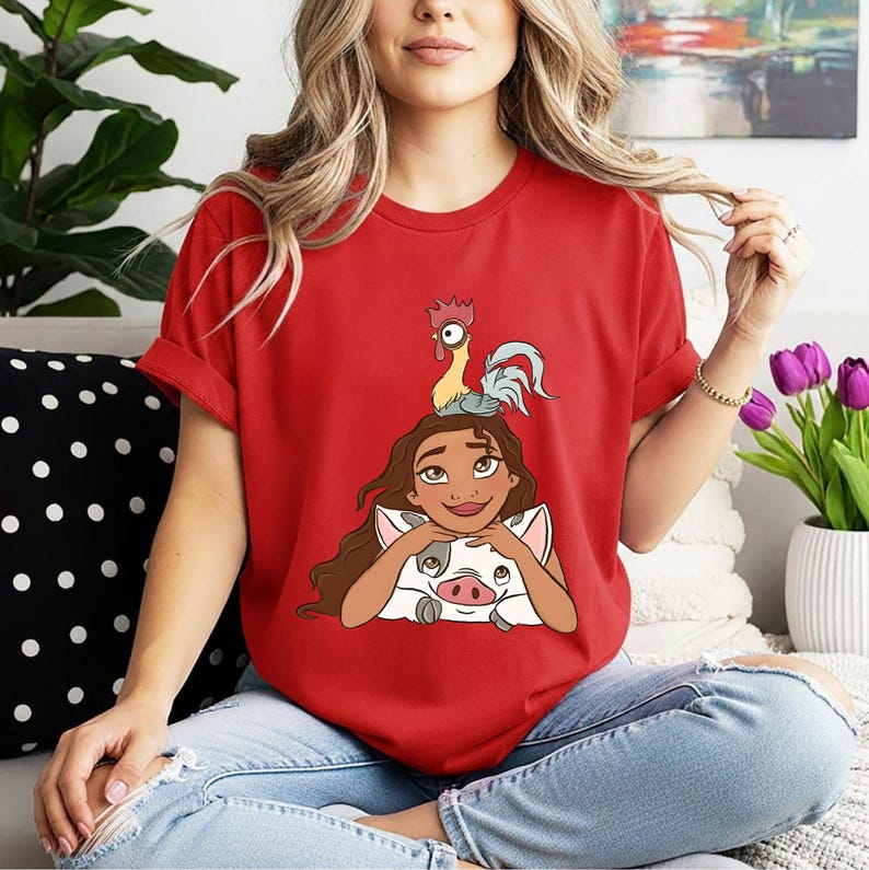 May include: A red t-shirt with a cartoon illustration of a character with long brown hair, a pig, and a rooster. The character is smiling and looking upwards. The shirt is made of a soft material.