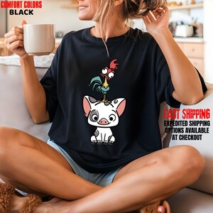 May include: Black t-shirt featuring a cartoon pig with a rooster on its head. The pig is white with black spots and pink ears. The rooster is red, blue, and yellow. The shirt has the words "COMFORT COLORS BLACK" and "FAST SHIPPING" printed on it.