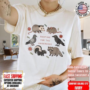 May include: An ivory short-sleeve t-shirt featuring a graphic print of an opossum, mouse, pigeon, crow, skunk, and raccoon arranged in a circle. The text "ROOTING FOR THE UNDERDOGS" is printed in the center in red letters, surrounded by small red stars.