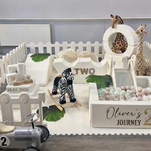 May include: A white play area with a fence, slide, and ball pit. Includes a toy car, rocking zebra, and stuffed giraffes. The play area is decorated with tropical leaves and the text "Oliver's Journey 2".