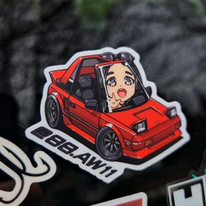 May include: A red sticker featuring a cartoon illustration of a red sports car with a person inside. The car has black wheels and the text "88.AW11". The person is waving with a surprised expression.