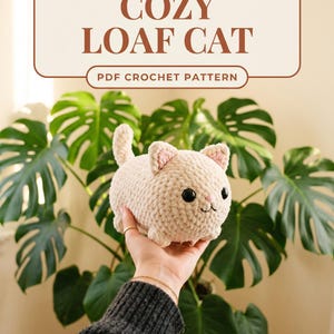 以下が含まれることがあります： 手で持たれた、パンの形をしたクリーム色の編み猫。 猫はピンク色の内耳と黒い目をしています。 「COZY LOAF CAT」と「PDF CROCHET PATTERN」の文字が猫の上に表示されています。