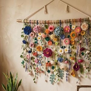 May include: A wall hanging featuring a horizontal wooden branch suspended by a cream-colored rope. Numerous colorful fabric flowers, including roses, daisies, and hydrangeas, are attached to hanging strings along with green leaves and vines.