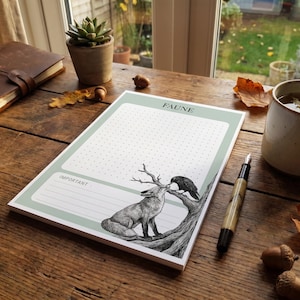 May include: A rectangular notepad with a sage green border, featuring a dotted grid section, a lined section labelled IMPORTANT, and a black and white illustration of a fox and a crow on a tree branch, with the word FAUNE printed at the top.
