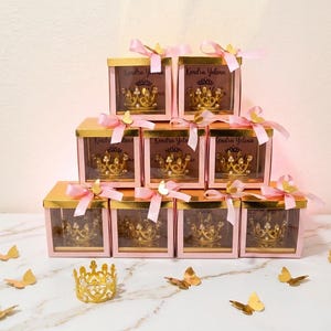 May include: A pyramid stack of nine square pink gift boxes with gold lids, each containing a gold crown with pearls. Each box is tied with a pink ribbon and a small gold butterfly. The text Kendra Yelena is printed on the boxes. A loose gold crown and several gold butterfly cutouts are arranged in front.