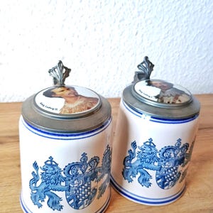 Peut inclure: Deux chopes à bière en céramique blanche ornées de motifs héraldiques bleus représentant des lions et des armoiries. Chaque chope possède un couvercle en métal surmonté d'une pastille en céramique avec le portrait du roi Louis II et l'inscription King Ludwig II.
