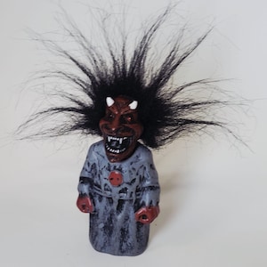 May include: A small figurine featuring a dark red, horned devil head with a wide, toothy grin and wild, spiky black hair. The figure wears a distressed, mottled grey robe with a circular red emblem on the chest and has dark red hands.