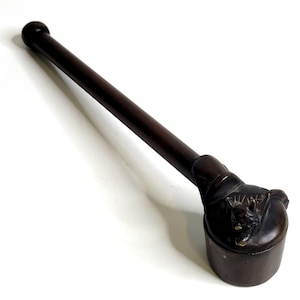 May include: A dark brown wooden pipe with a long, slender stem and a rounded bowl. The bowl features a carved, detailed animal figure on its top surface, with a smooth, polished finish throughout the entire object.