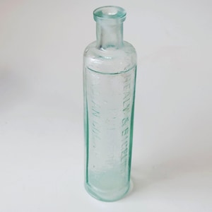 May include: A clear, light aqua-tinted glass bottle with a cylindrical body and a narrow neck. The glass features embossed text that reads HEALY & BIGELOW and INDIAN COUGH CURE, with a rounded rim at the top and a smooth, slightly reflective surface.