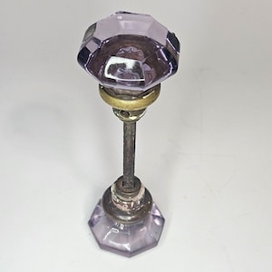 May include: A vintage door knob with a faceted, light purple glass top and bottom. The knob has a brass collar and a threaded metal rod. The knob is likely from an antique door.