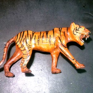 May include: A handcrafted tiger figurine featuring a glossy orange finish with distinct black stripes. The tiger is depicted in a walking stance with its mouth open, revealing white teeth and a yellow eye, capturing a dynamic and alert posture.