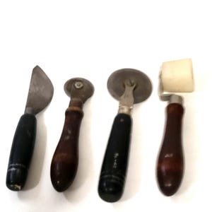 Puede incluir: Un juego de cuatro utensilios de cocina que incluye una hoja de metal curva con mango negro, una rueda de metal pequeña con mango de madera oscura, una rueda de metal más grande con mango negro marcado No. 40 y un rodillo de plástico blanco.