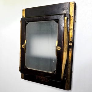 May include: A vintage dark wood and brass camera ground glass focusing screen frame. The rectangular device features a central frosted glass panel held by brass clips and side levers, with a small metal plate at the bottom edge.