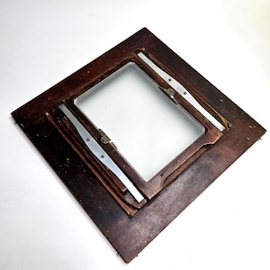May include: A square dark brown wooden camera film holder frame featuring a central rectangular opening. The frame includes silver-toned metal sliding tracks and small metal fasteners attached to the wood, showing a vintage construction.