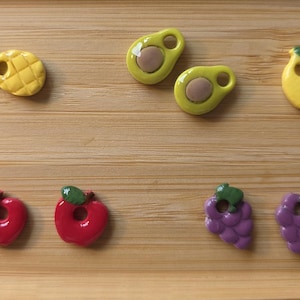 Peut inclure: Un ensemble de dix petites breloques en forme de fruits disposées par paires. Le lot comprend des ananas jaunes, des avocats verts et jaunes, des citrons jaunes, des pommes rouges et des raisins violets. Chaque breloque a un petit trou circulaire.