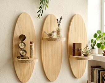 May include: Three oval-shaped, light-colored wooden shelves mounted on a cream-colored wall. Each shelf holds decorative items: a compass, a small bird figurine, a leather-bound journal, and a glass terrarium. Green plants hang above.