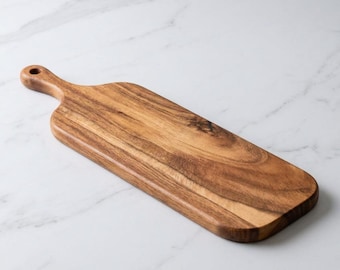 May include: A rectangular wooden cutting board with a handle and a hole for hanging. The board is made of a light brown wood with a natural grain pattern. The handle is rounded and smooth. The board is on a white marble surface.