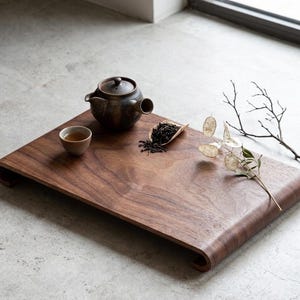 May include: A wooden tea tray with a curved edge, holding a small ceramic teapot, a cup of tea, and a wooden scoop of loose tea leaves. Dried decorative branches and leaves are arranged on the tray. The tray is a rich brown color.