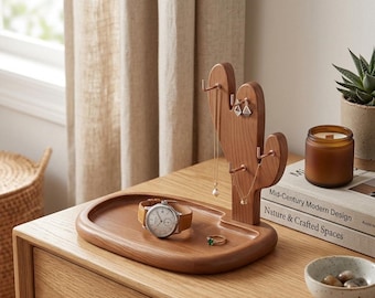 May include: A wooden cactus-shaped jewelry holder with a tray, holding a watch, ring, and necklaces. A candle and a small bowl with stones are also on the wooden surface. The jewelry holder has hooks for earrings and necklaces.