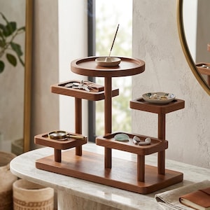 May include: A multi-tiered wooden organizer with various trays and platforms. The organizer holds keys, a magnifying glass, jewelry, and other small items. An incense stick burns on the top tier. The organizer is made of a warm-toned wood.