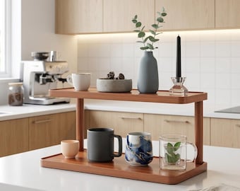 May include: A two-tiered wooden serving tray displays various kitchen items. The top shelf holds a white mug, a small concrete bowl with decorative items, a blue vase with greenery, and a black candle. The bottom shelf has mugs and a glass cup.