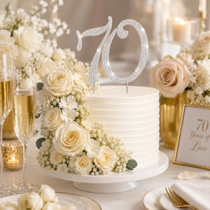 May include: A silver-toned cake topper shaped as the number 70, covered in sparkling rhinestones. The topper is positioned on top of a white, horizontally textured cake decorated with a cluster of cream-colored roses and small white flowers.