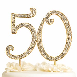 May include: A gold-colored cake topper in the shape of the number 50, encrusted with sparkling rhinestones. The numbers are mounted on gold-colored stems and are placed above white flowers. This festive decoration is perfect for a milestone birthday celebration.