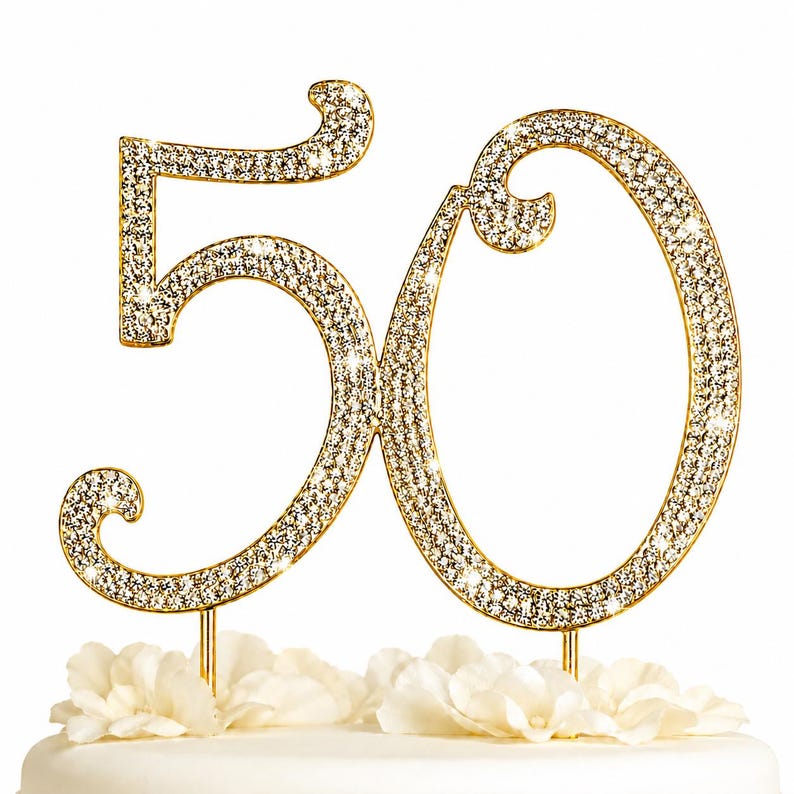 May include: A gold-colored cake topper in the shape of the number 50, encrusted with sparkling rhinestones. The numbers are mounted on gold-colored stems and are placed above white flowers. This festive decoration is perfect for a milestone birthday celebration.