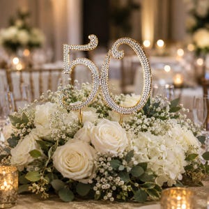 May include: A gold-colored, rhinestone-studded number 50 cake topper. The numbers are set atop a bed of white roses, hydrangeas, and greenery. The arrangement is on a table with candles and other floral arrangements.