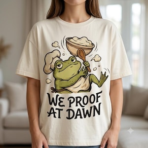 May include: A cream-colored t-shirt featuring a graphic of a green frog wearing a chef hat and apron, holding a wooden spoon and a bowl of dough. Below the illustration, the text reads "WE PROOF AT DAWN" in bold, black, stylized lettering.