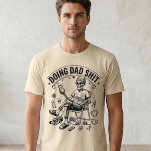 Könnte beinhalten: Ein beiges Kurzarm-T-Shirt mit einer schwarzen Grafik eines Skeletts, das in einem Klappstuhl sitzt und ein Telefon sowie einen Becher hält. Der Text "DOING DAD SHIT" steht darüber, umgeben von Illustrationen von Babyartikeln und Werkzeugen.