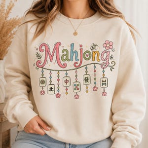 May include: A cream-colored crewneck sweatshirt featuring the word Mahjong in colorful, stylized lettering. Below the text, a decorative string hangs with various mahjong tiles, small colorful beads, and pink flower accents.