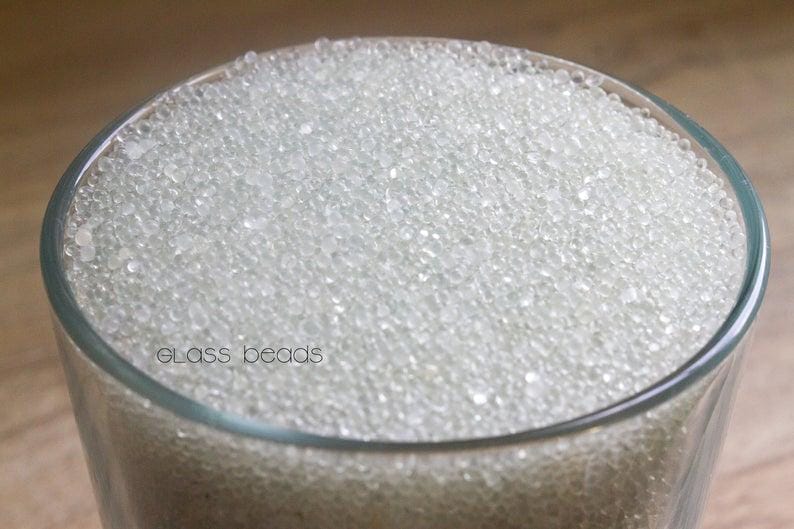 May include: A clear glass jar filled with small, white glass beads. The jar is sitting on a wooden surface. "glass beads" is written on the jar.