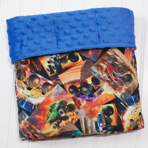 May include: A blue and patterned blanket featuring monster trucks in action. The top portion is a solid blue, textured fabric. The lower portion has a patchwork design with images of trucks in various colors, set against a fiery background.