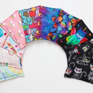 May include: A set of nine colorful fabric heat packs with various designs, including rainbows, candy, butterflies, and cats. The heat packs are arranged in a semi-circle on a white wooden surface.