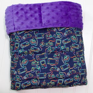 May include: A folded blanket featuring a dark blue fabric with a colorful pattern of various video game controllers and handheld consoles. The top section of the blanket is made of a solid purple textured minky fabric with raised dots.