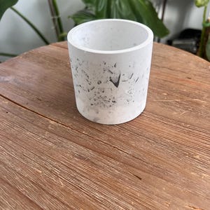 May include: A cylindrical, white concrete planter with a speckled pattern of dark gray and black. The planter is open at the top and sits on a wooden surface. The background includes green plants.