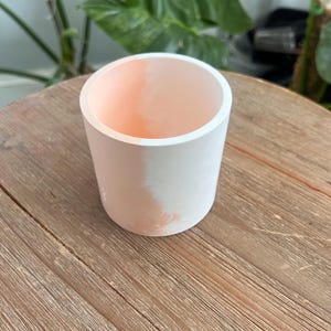 May include: A small, cylindrical planter with a white exterior and a peach-colored interior. The planter is sitting on a wooden surface. The interior color is visible at the top of the planter.