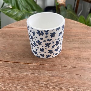 May include: A white ceramic plant pot with a blue floral pattern. The cylindrical pot has a smooth, glossy finish and is sitting on a wooden surface. The blue floral design is evenly spaced around the exterior of the pot.